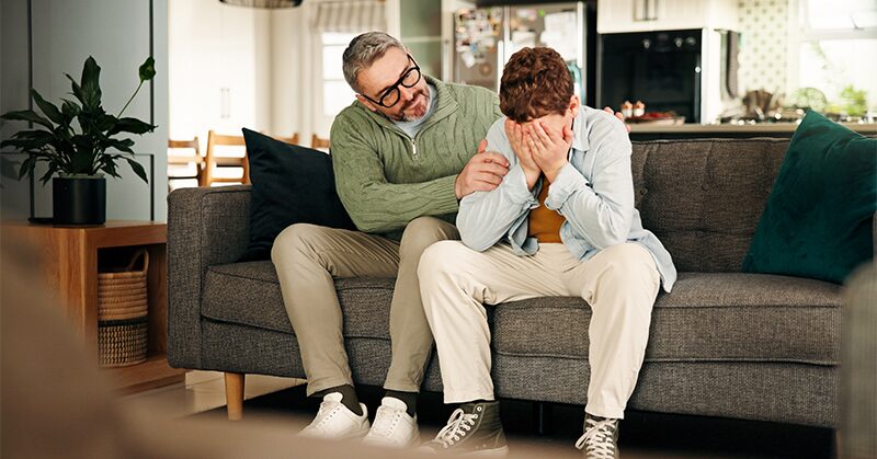 A parent and teenager on a couch in a family session, the teenager covering their face.