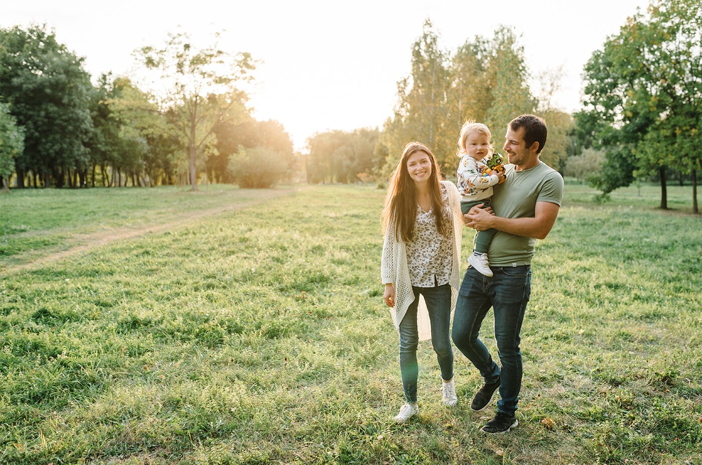 A family of three walking together across an open sunlit park at golden hour.
