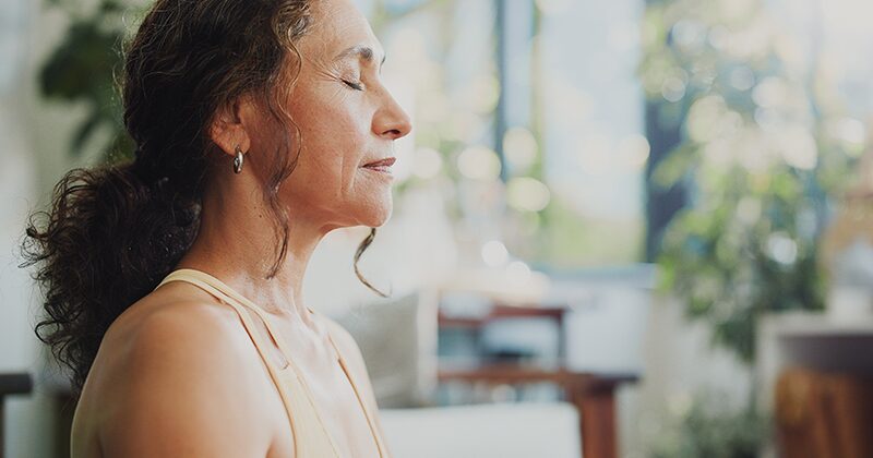 A woman sitting quietly with her eyes closed in a bright room, taking a slow breath.