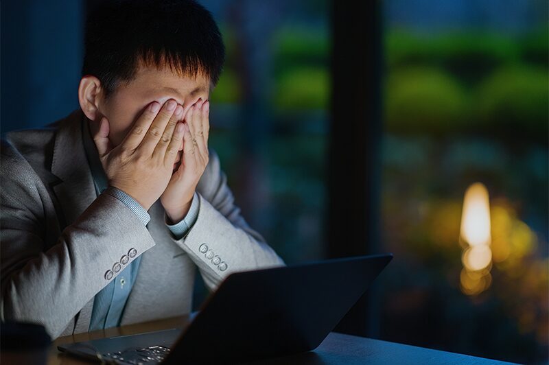 A man working late at a laptop, resting his face in his hands.