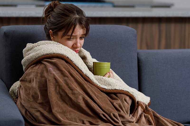 A woman wrapped in a blanket on a sofa, holding a warm mug with both hands.